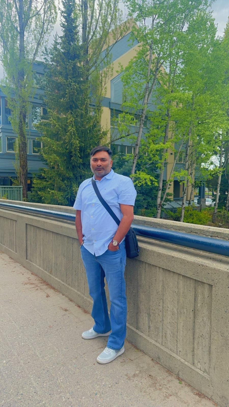 Man in casual attire standing on a concrete bridge with a forested building in the background.