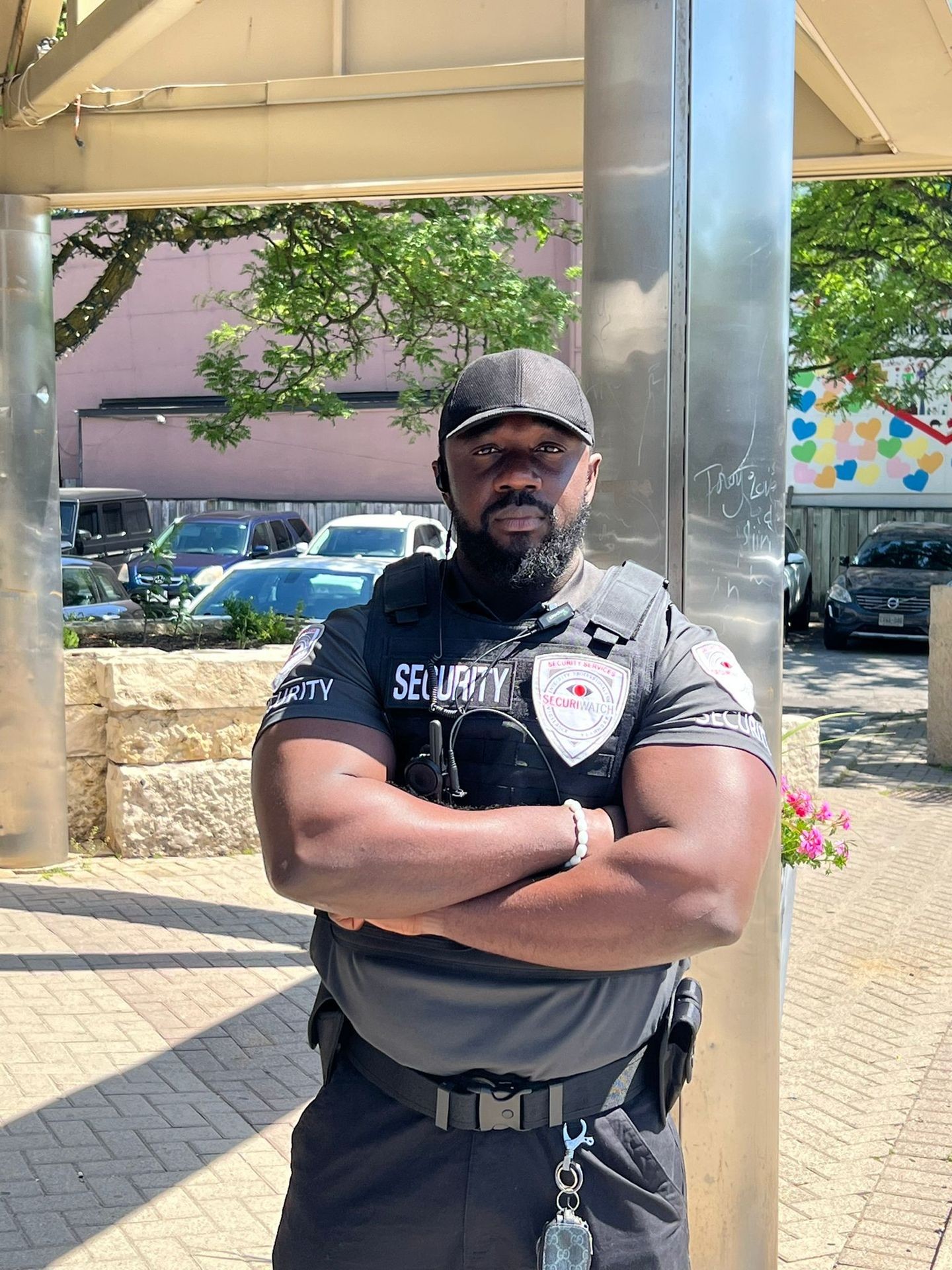 Security guard in uniform standing with arms crossed in front of a parking lot and building.