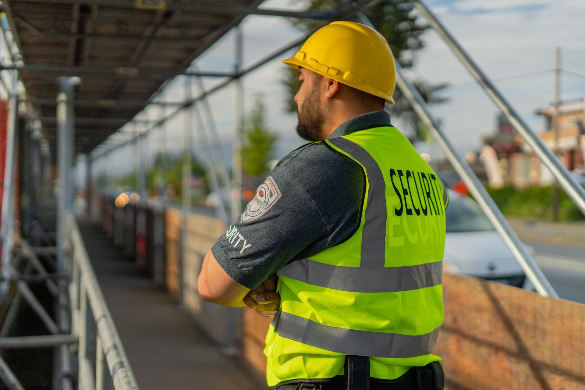 Security guard in a yellow hard hat and reflective vest standing on a construction site with scaffolding.