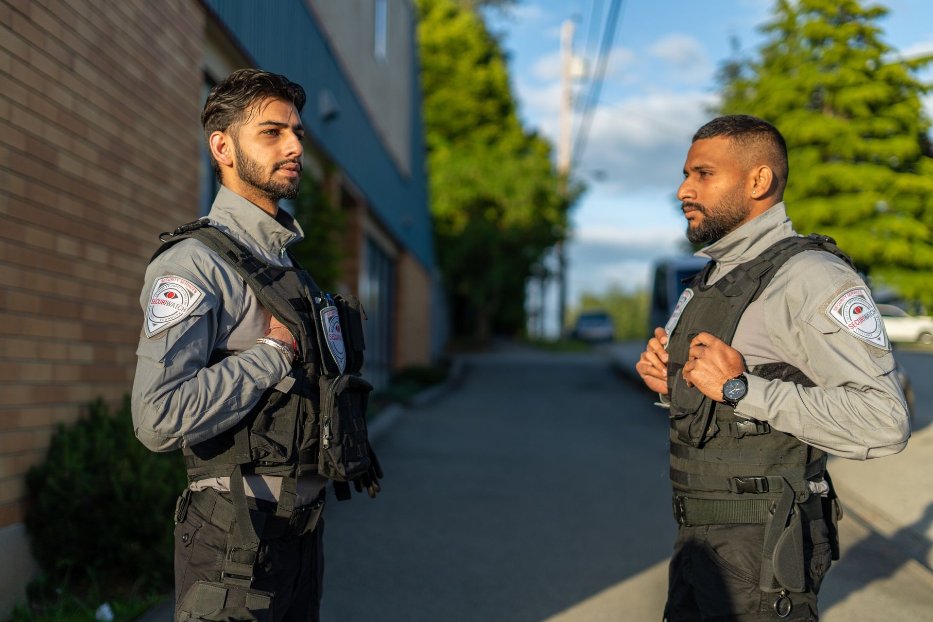 Two security personnel wearing vests standing outdoors on a street with trees and buildings in the background.