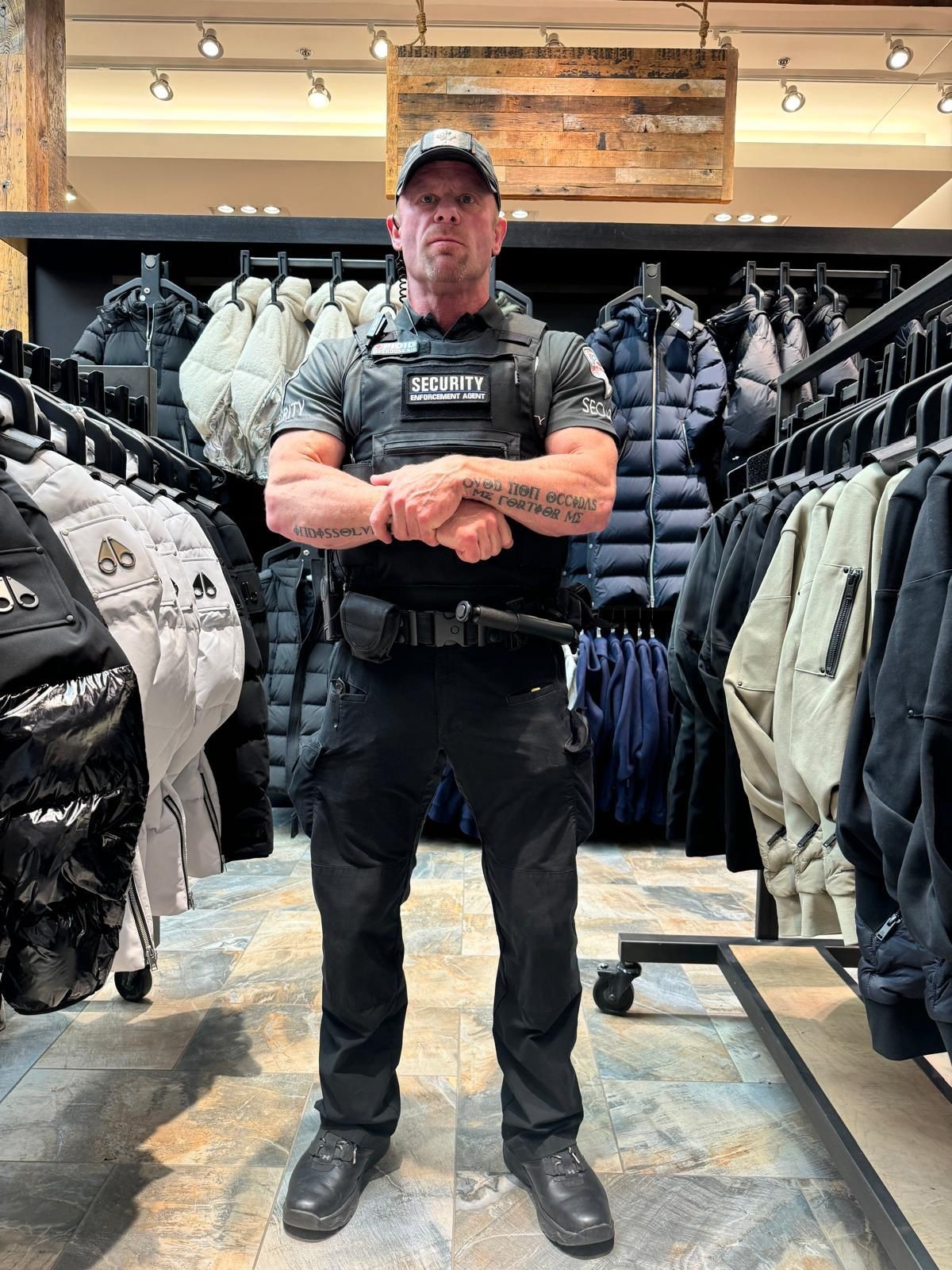 Security guard in uniform standing with crossed arms in a clothing store, surrounded by racks of jackets.