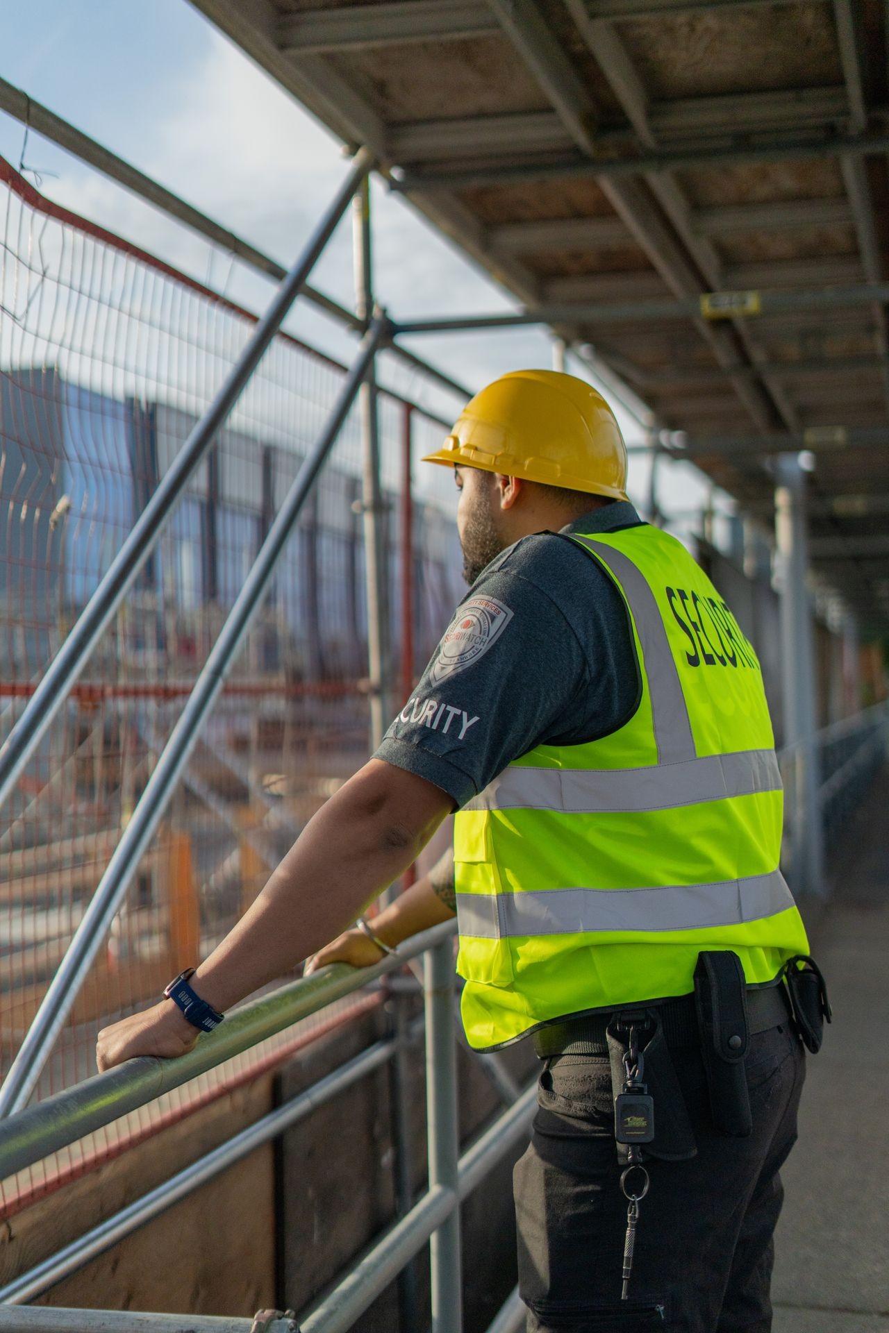 Security guard wearing a hard hat and reflective vest observes a construction site from behind a safety barrier.