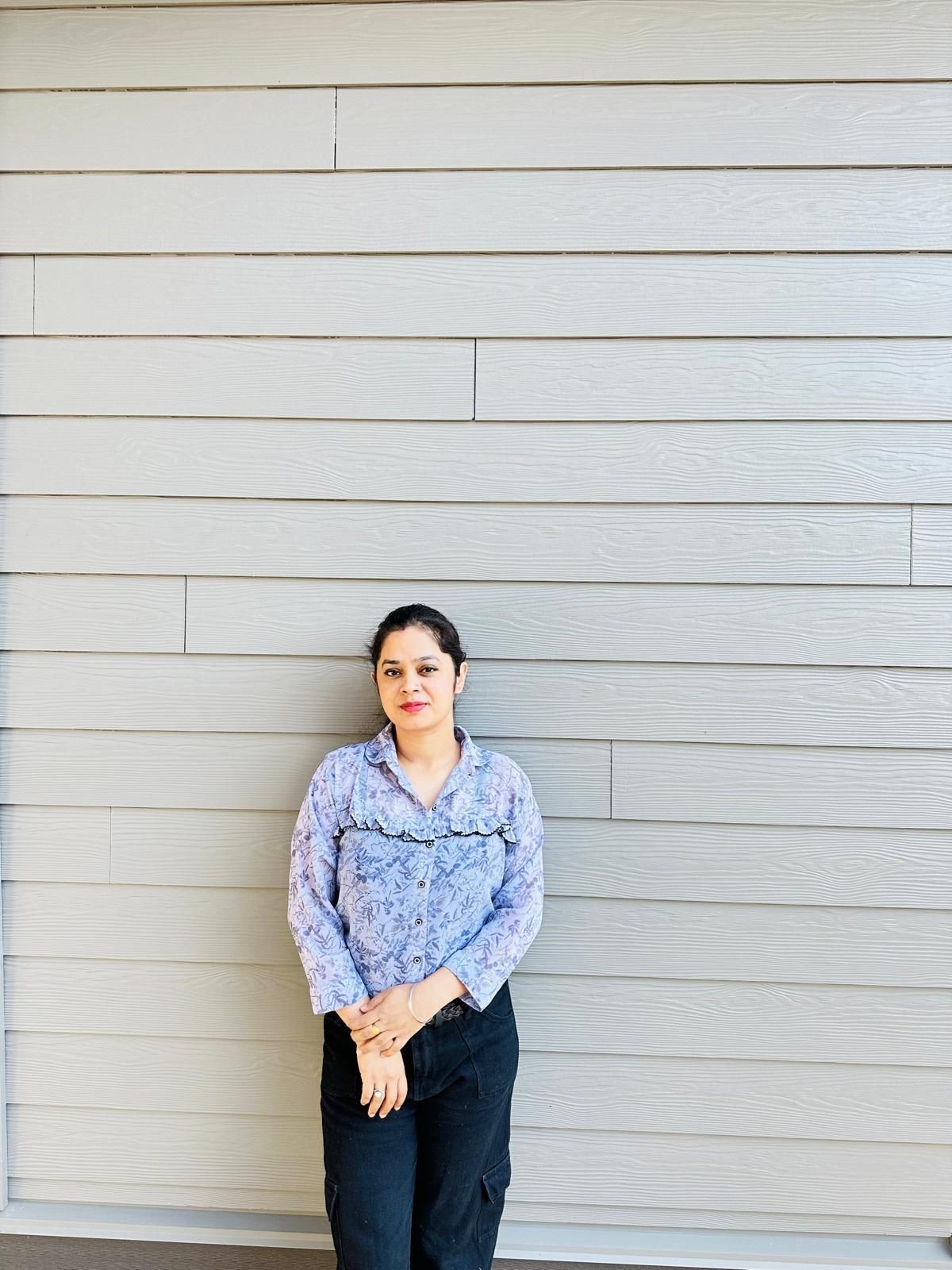 Person standing in front of a light gray wooden wall, wearing a patterned top and dark pants.