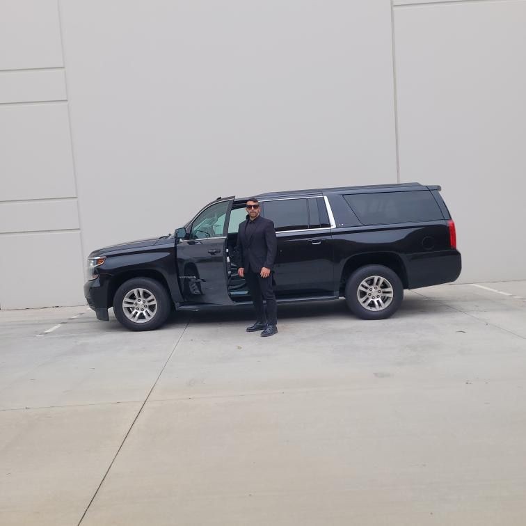 Man in black suit and sunglasses standing next to a large black SUV with the driver's door open.