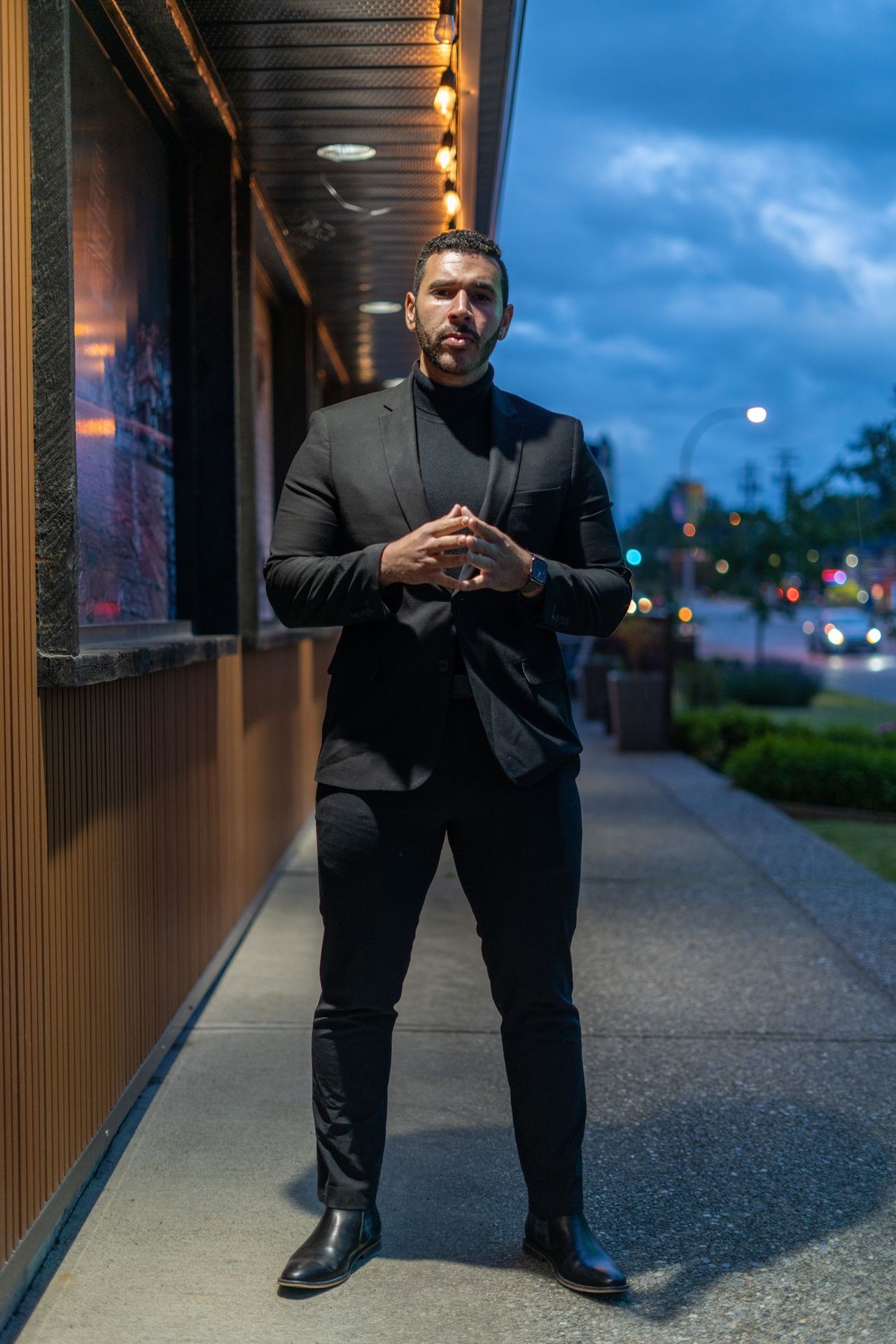 Individual wearing a black suit and boots, standing on a sidewalk with evening lights illuminating the street.