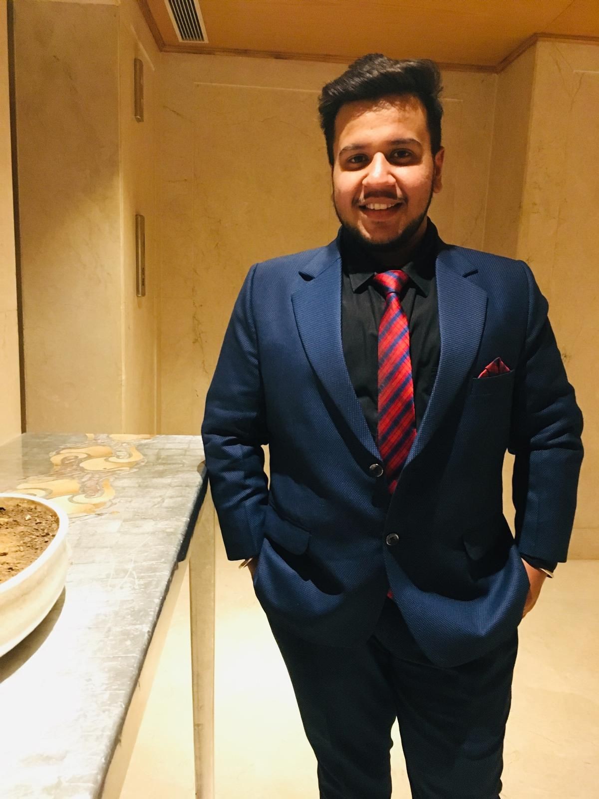 Smiling man in a blue suit and red striped tie standing indoors with hands in pockets.