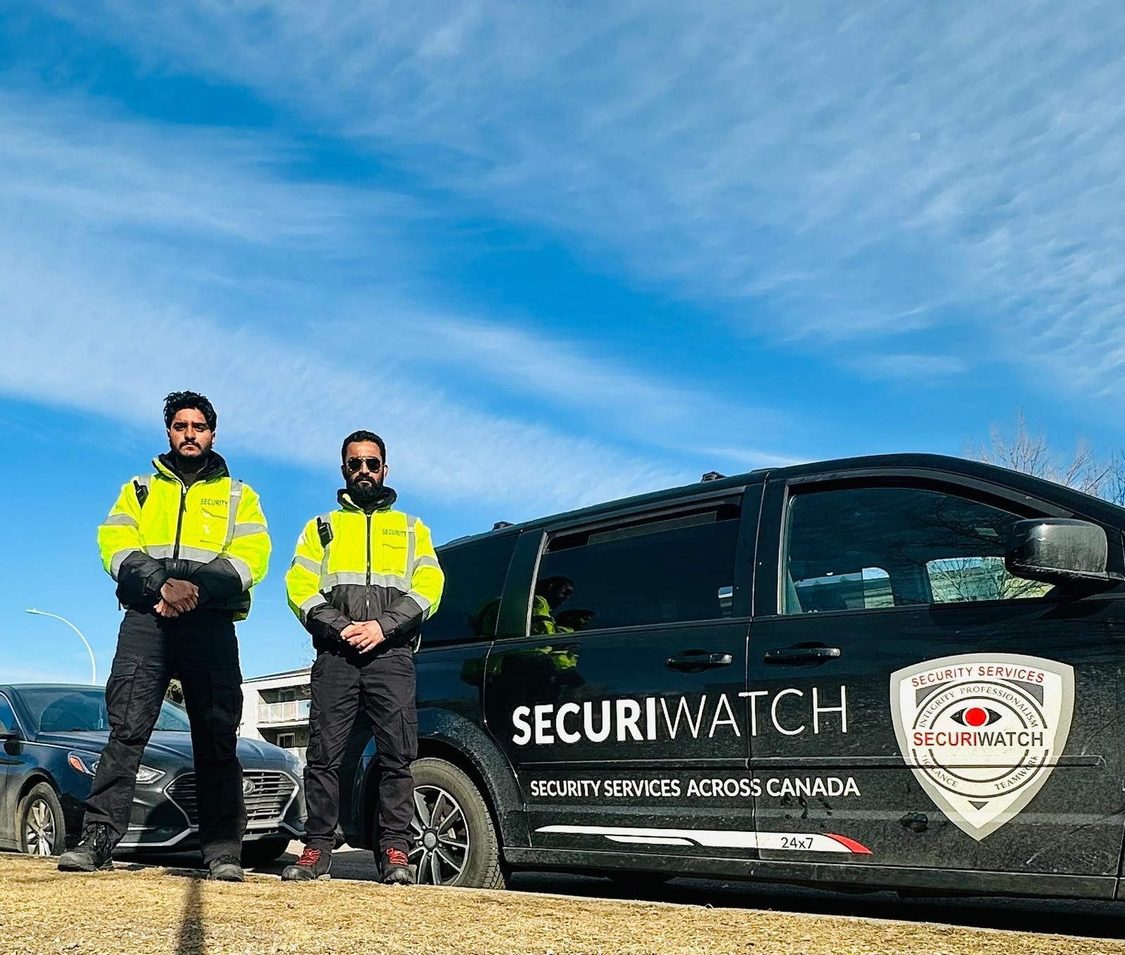 Two security guards in yellow jackets standing next to a SecuriWatch vehicle on a sunny day.