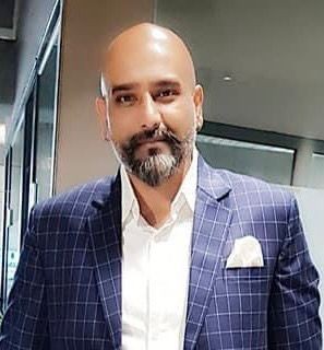 Man with a beard wearing a checked suit jacket and a white shirt, standing indoors.