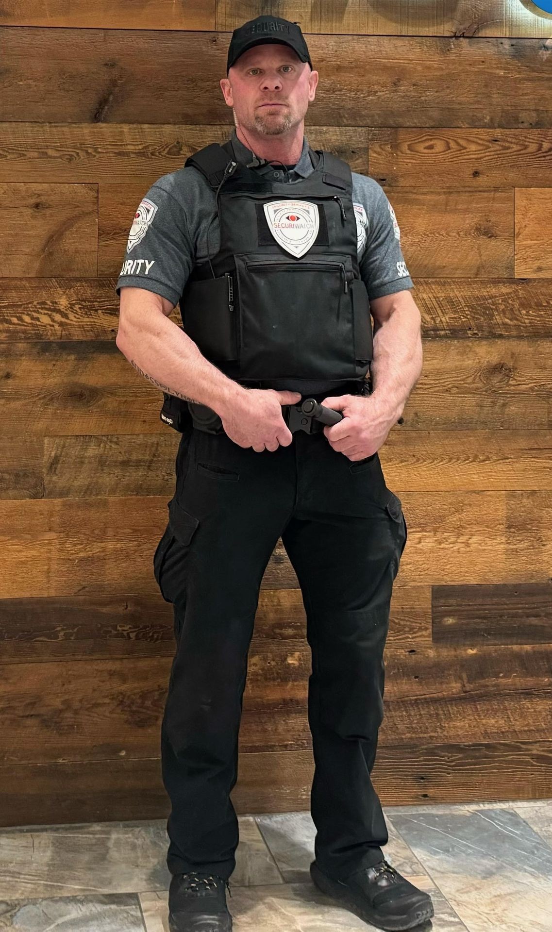 Security guard in uniform and body armor standing against a wooden wall, holding a security baton.