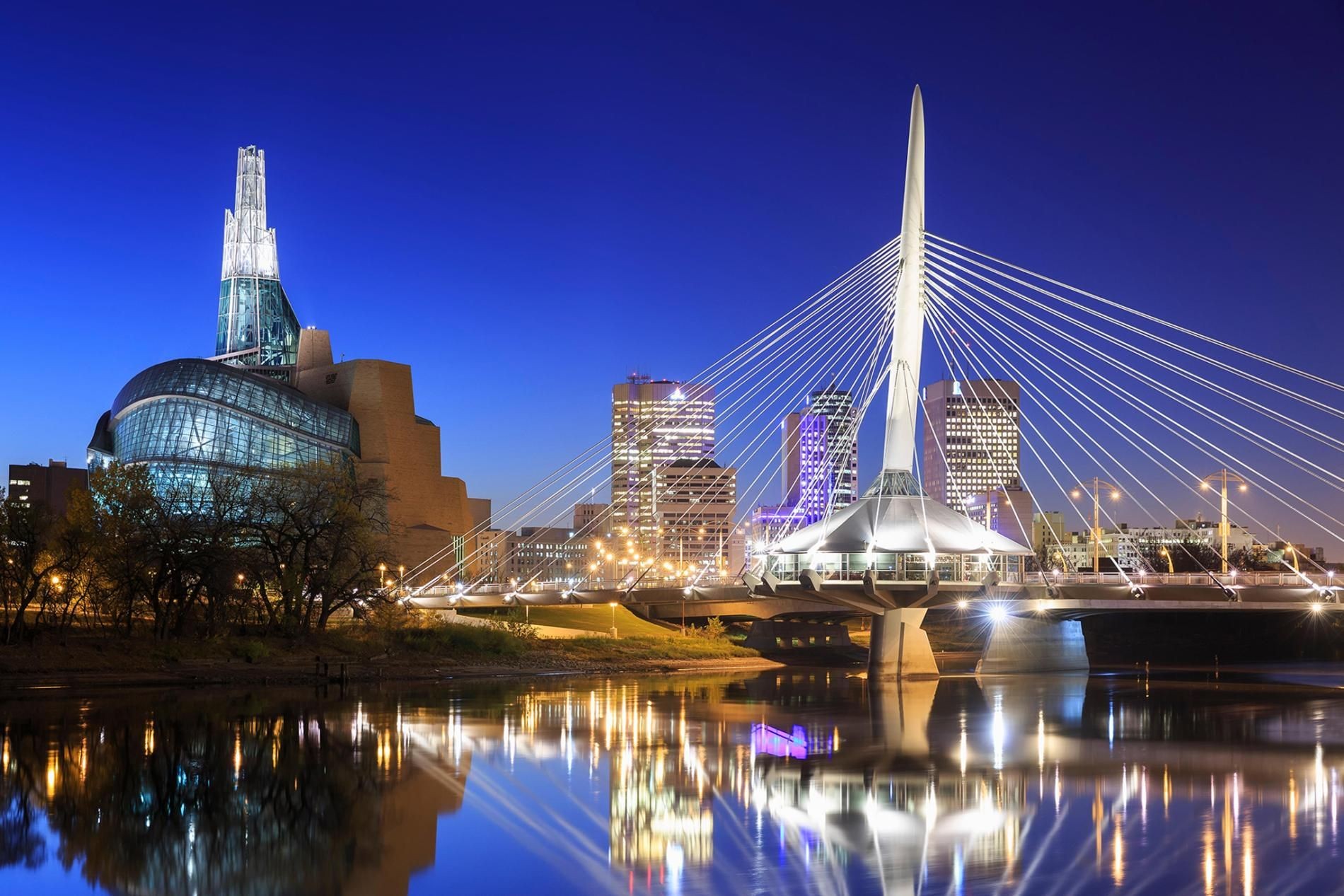 Night view of a modern bridge and illuminated buildings reflecting on the water in a cityscape.