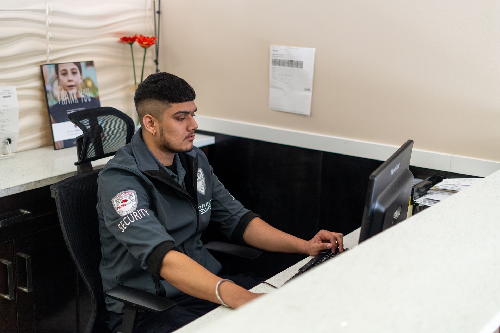 Security personnel seated at a desk using a computer, with office decor and documents in the background.